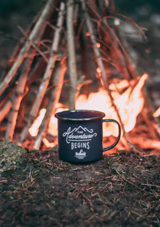 A cozy coffe cup that says The Adventure Begins, next to a warm fire.